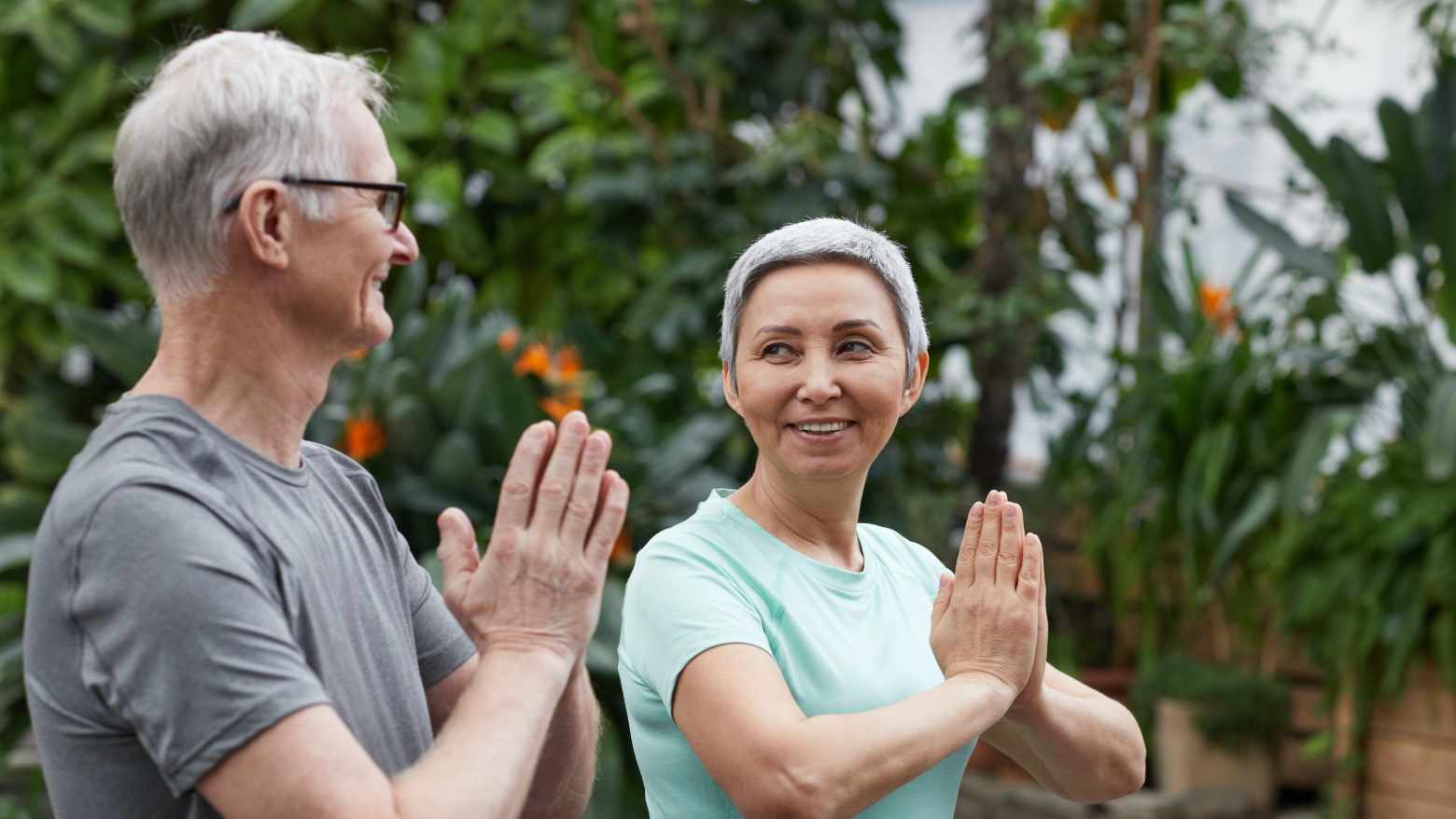 An elderly husband and wife doing yoga poses in a garden centre. 