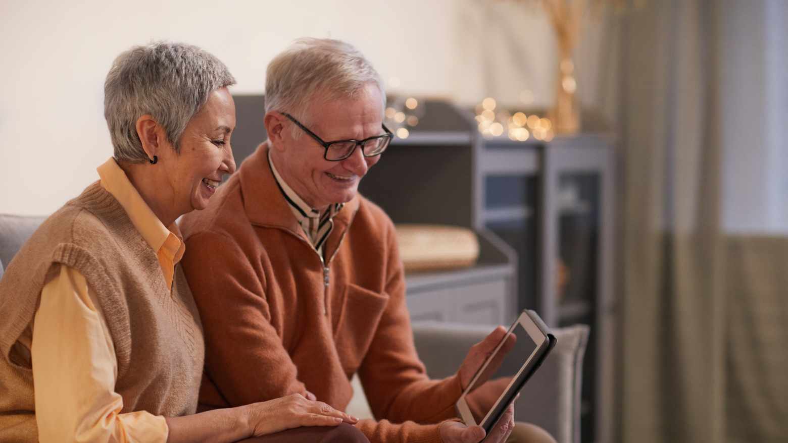 An elderly husband and wife sitting on a sofa smiling at a tablet device. 