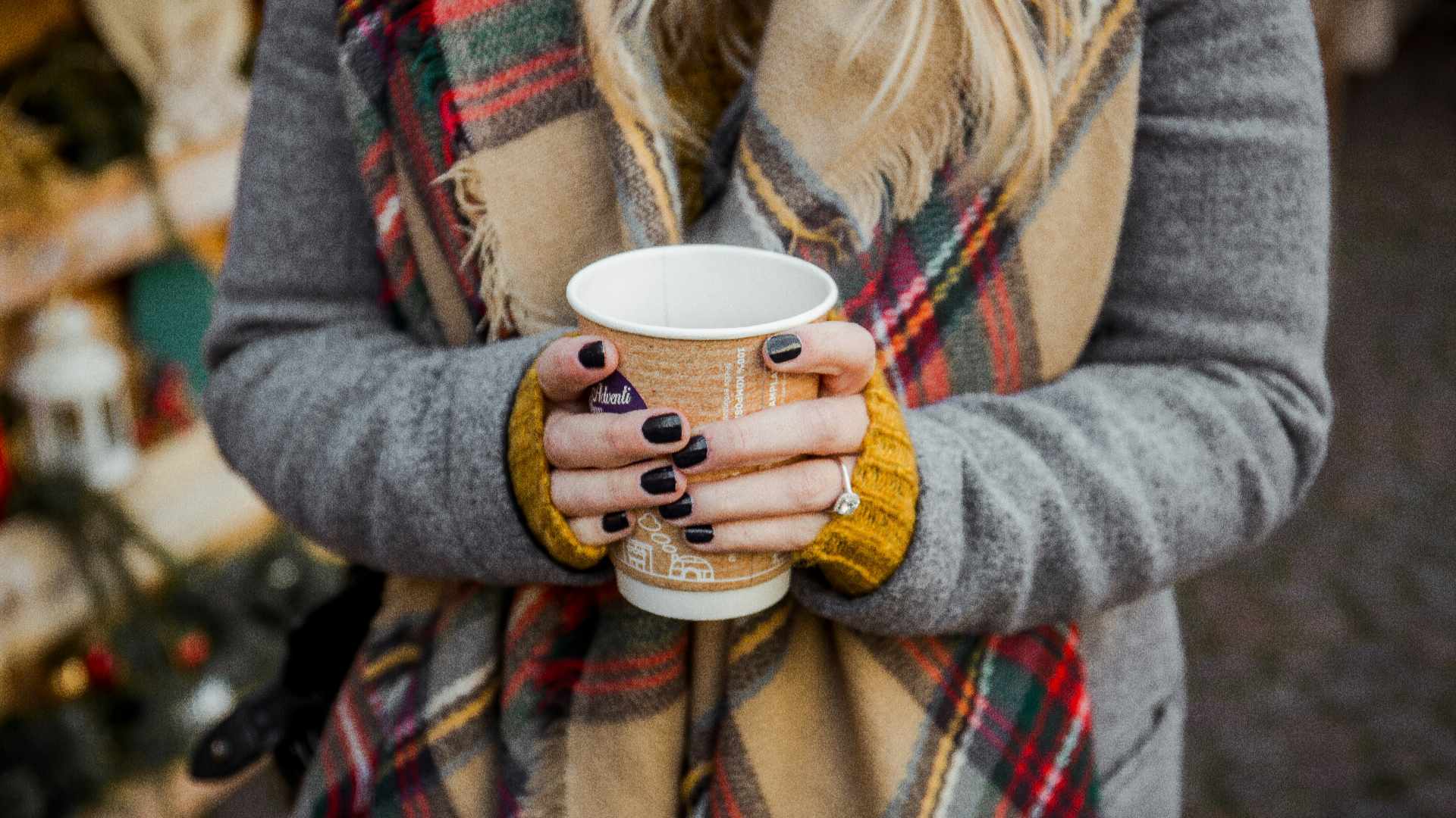 A lady in a grey coat and yellow jumper and tartan scarf holding a coffee cup with two hands outside at a Christmas market