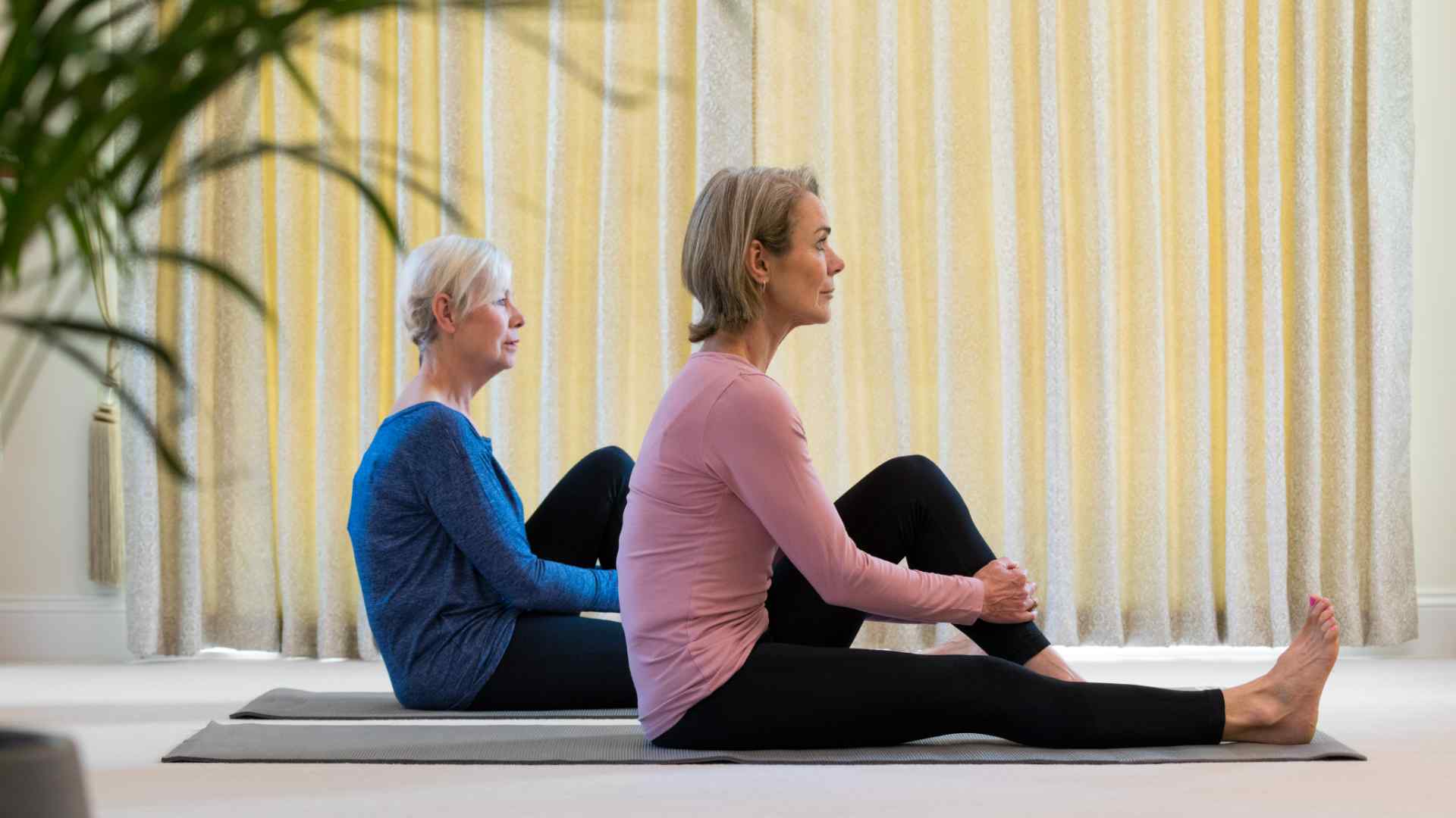 Two elderly ladies doing yoga inside. One in a pink top and leggins, one in a blue top and leggins.