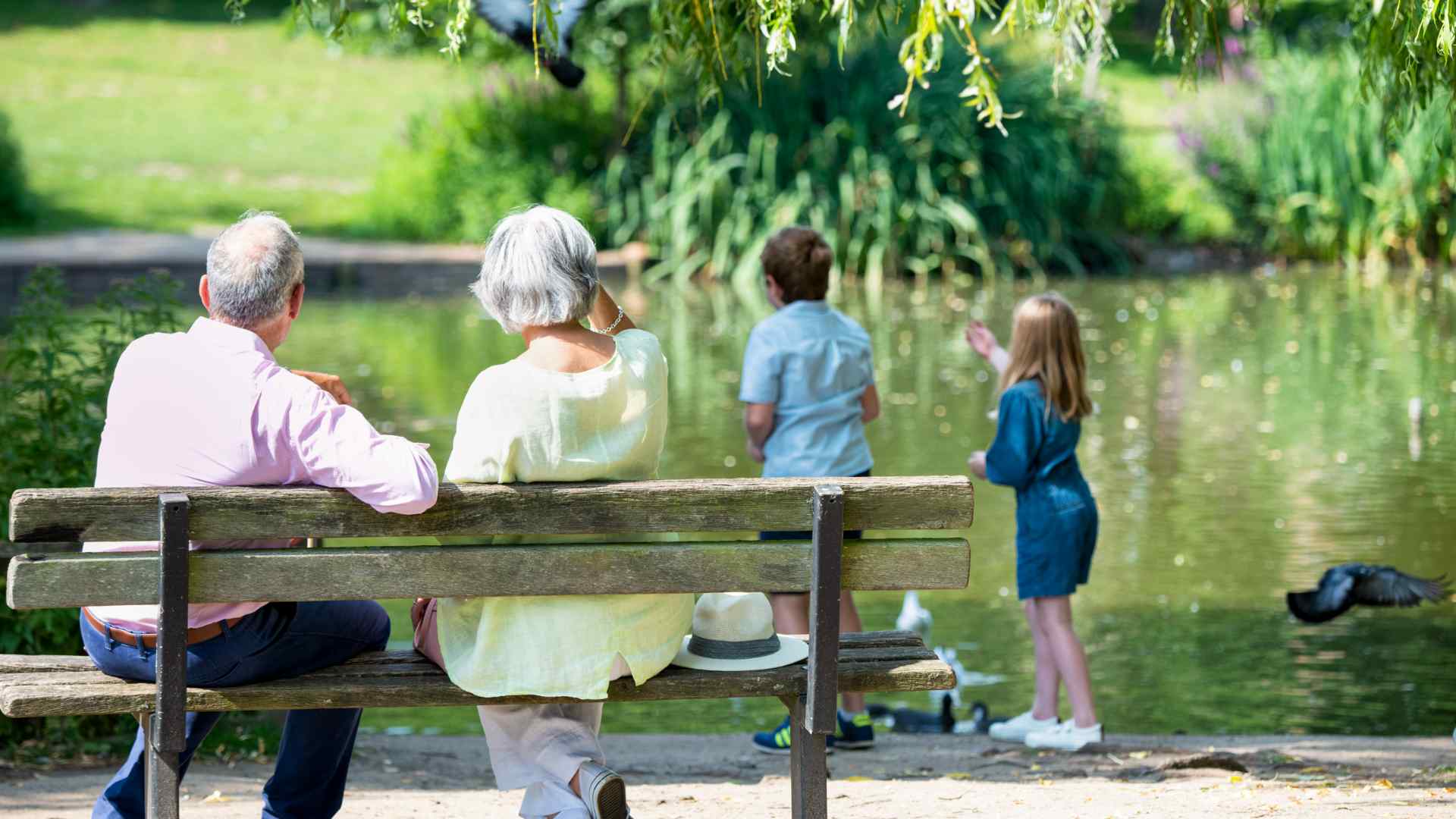 Two grandparents watching their grandchildren feed the ducks.