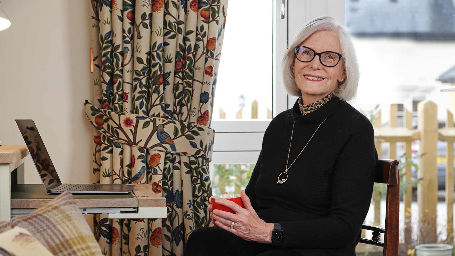 Jane Reyanrd in a black outfit drinking coffee from a red mug on a laptop by her desk