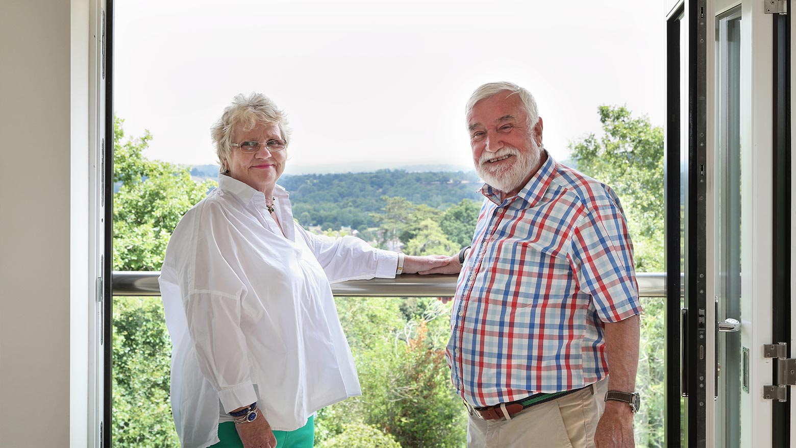A husband and wife standing together on a balcony in a stylish apartment with trees in the background.