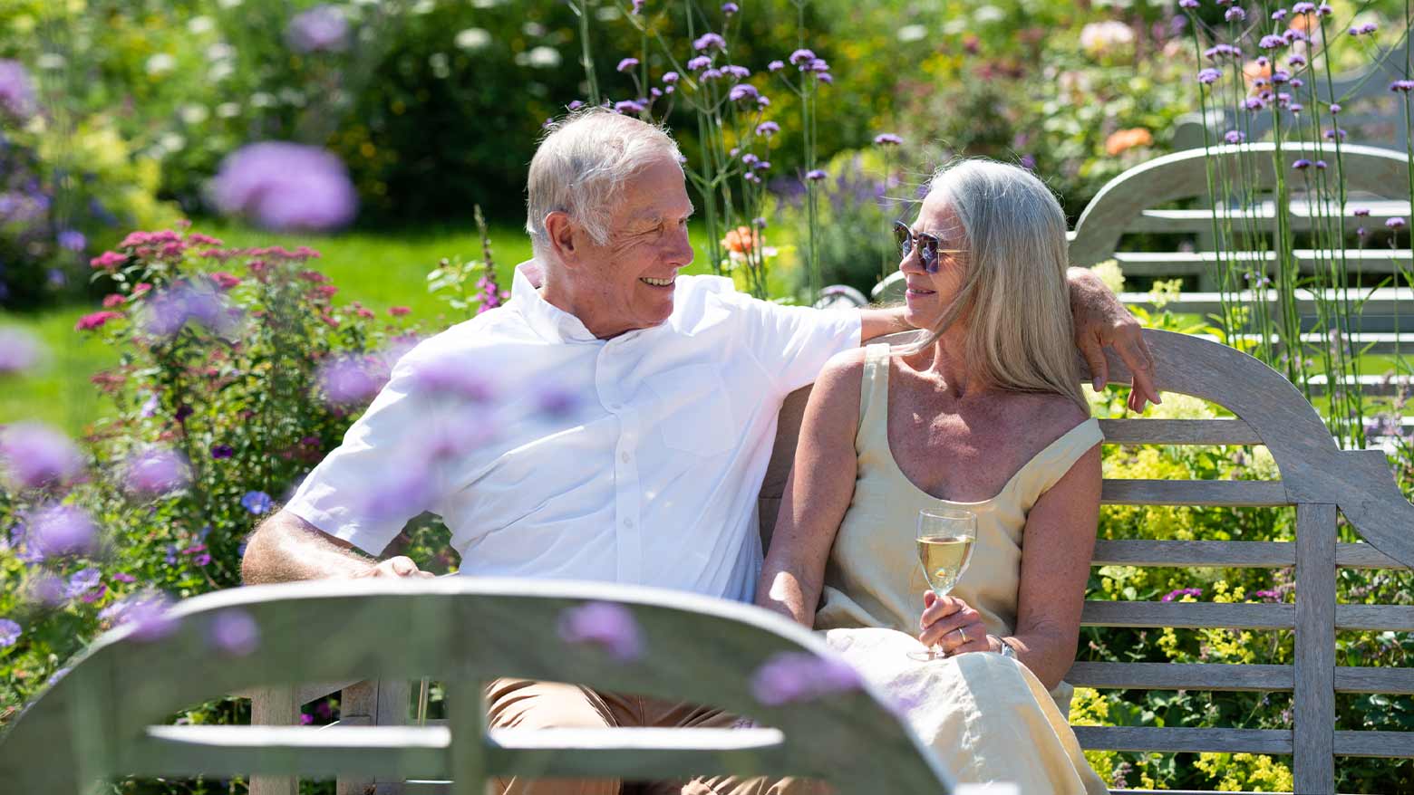 An older couple sat on a bench in a beautiful flowered garden. The lady looks into the mans eyes holding a glass of white wine.