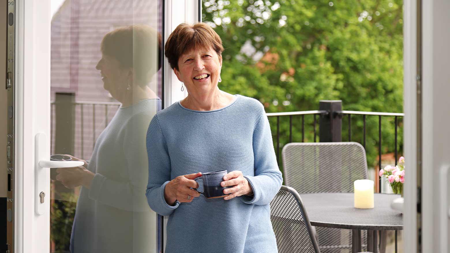 An older lady in a blue jumper holding a blue mug of coffee next to a balcony